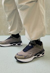 Sneakers i grått och olivgrönt, med en slät textilöverdel, kontrasterande marinblå snören och en vit sula med luftdämpningsinslag.