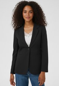 Blazer noir au design ajusté, à bouton unique, avec des revers crantés et deux poches avant, porté sur un haut blanc orné de dentelle.