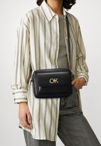 Borsa a tracolla in pelle nera con logo dorato, forma rettangolare e chiusura con zip, indossata sopra una camicia a righe e pantaloni grigi.