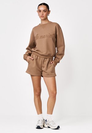 Młoda kobieta w brązowej bluzie z długim rękawem i pasujących krótkich spodenkach z kieszeniami, białych skarpetkach i białych sportowych butach, stoi na białym tle.