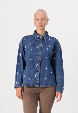 Denim jacket, blue with white floral embroidery. Features a button-up front, pointed collar, and loose fit. Soft texture and casual style.