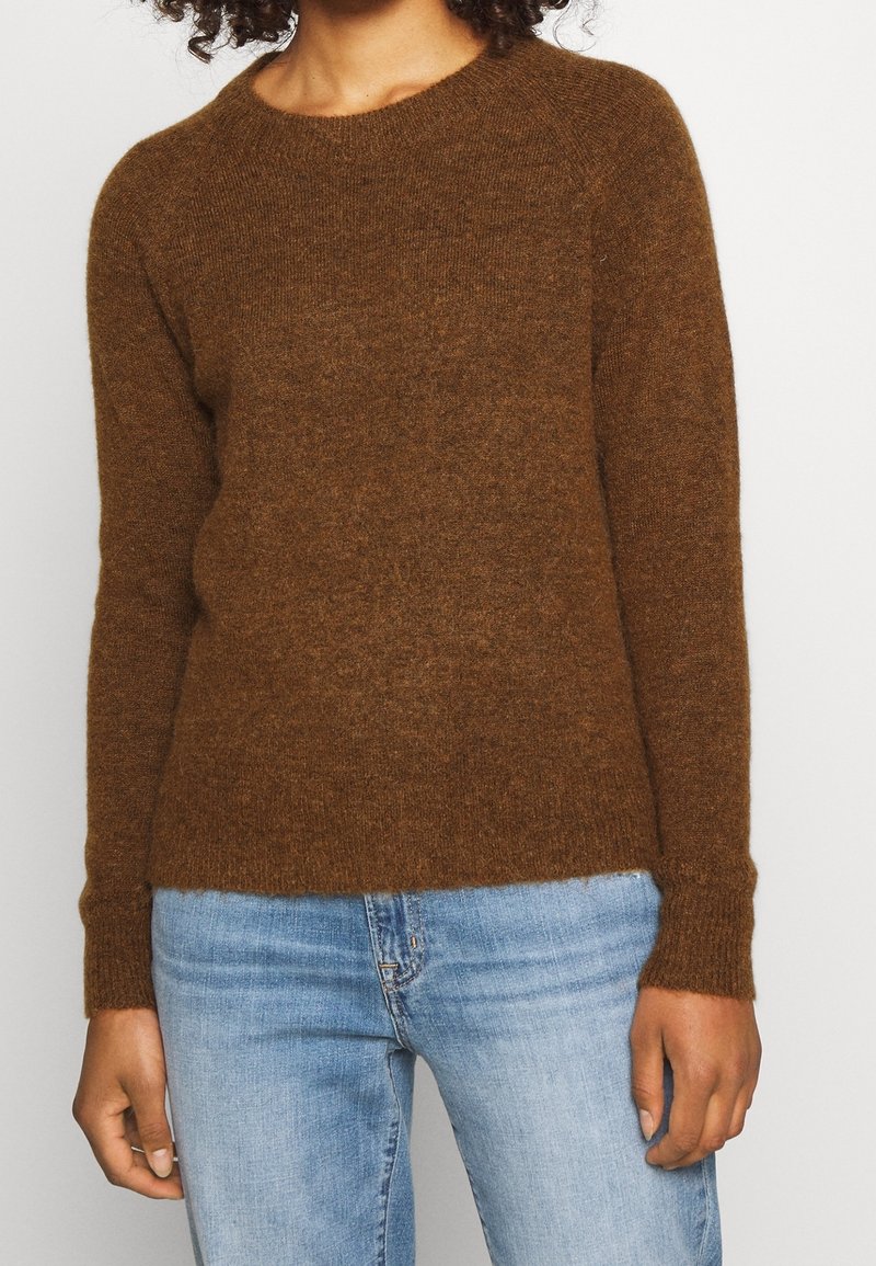 Pull marron tricoté avec un col rond, des manches longues et des poignets côtelés. Texture douce et coupe décontractée. Porté avec un jean bleu clair.