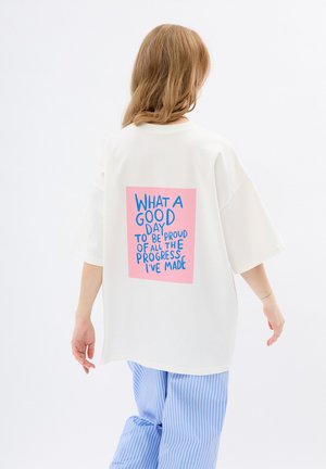 Witte oversized t-shirt met korte mouwen, voorzien van een roze rechthoekige grafiek op de achterkant met blauwe tekst, gedragen met blauw gestreepte broeken.