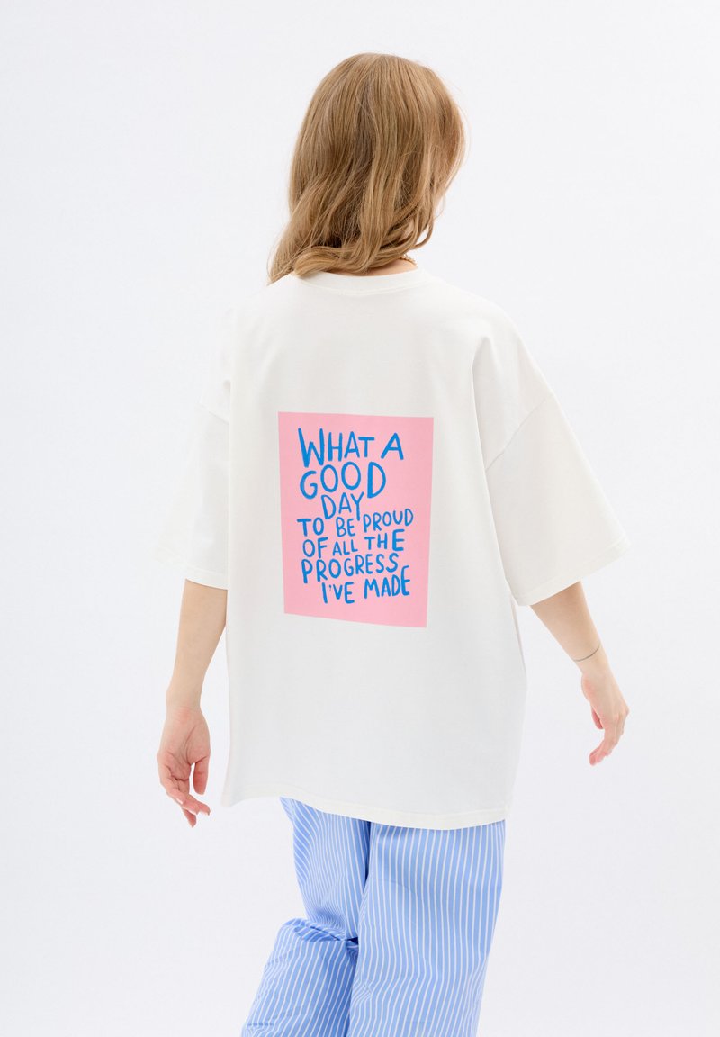 T-shirt blanche oversize à manches courtes, avec un graphisme rectangulaire rose au dos avec du texte bleu, portée avec un pantalon à rayures bleues.