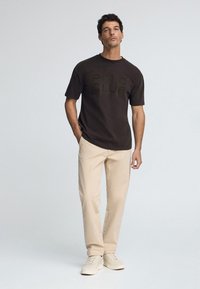 Brun bomullströja med präglad logo "POLO CLUB", kombinerad med gräddfärgade chinos. Outfiten fulländas med ljusa sneakers.