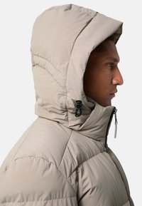 Man in een beige gewatteerde jas met de capuchon omhoog, getoond in rechterzijprofiel tegen een effen lichte achtergrond.