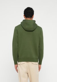 Zöld kapucnis pulóver, fűzős kapucnival, bordázott mandzsettákkal és aljjal. Sima textúra, laza dizájn, világos bézs nadrággal viselve.