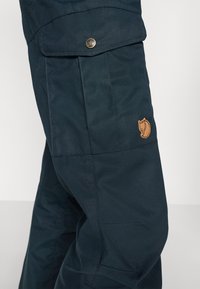 Marineblå cargobukser laget av slitesterkt stoff, med sydd detaljer, en sidelomme og en liten brun logopatch.