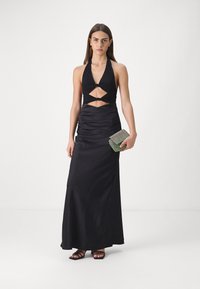 Rochie neagră din satin, cu gât halter și detalii tăiate la talie, având o siluetă ajustată și fustă fluidă. Modelul ține în mână o plică verde.