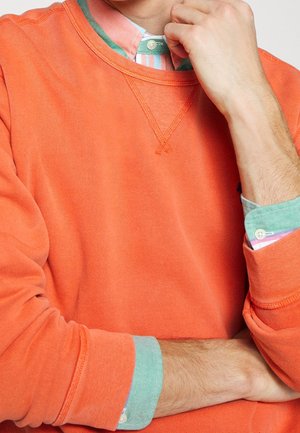 Uomo che indossa una felpa arancione sopra una camicia a righe verdi e rosa con bottoni, poggiando il mento sulla mano con il braccio incrociato.