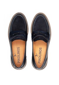 Mocasines azul marino hechos de piel suave con un diseño cosido y una planta acolchada en color marrón claro, que presenta el logo de la marca en el interior.