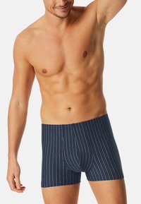 Uomo senza maglietta che indossa boxer blu navy con sottili righe verticali azzurre e bianche, con un braccio alzato sopra la testa.