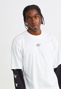 Tricou alb cu mânecă scurtă, guler rotund și un design grafic negru cu stele pe mânecile lungi. Logo mic pe piept.