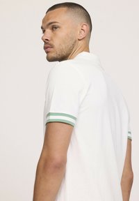 T-shirt polo blanc avec des poignets à rayures vertes, doté d'un col classique et de manches courtes. Le matériau semble lisse et léger.