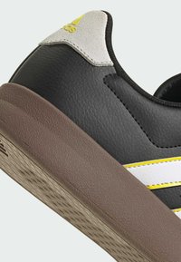 Crne kožne tenisice s smeškastom teksturiranom petom, bijelim i žutim trakama te sivim suedenim detaljem na peti. Imaju dizajn s niskim profilom.