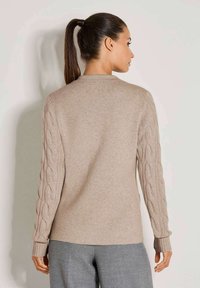 Frau mit einem dunklen Pferdeschwanz, die einen beigen Pullover mit Zopfmusterärmeln und grauen Hosen trägt, mit dem Rücken zu einer schlichten Hintergrundfläche.