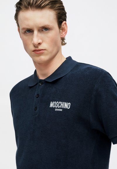 Νεαρός άνδρας που φοράει ένα μπλε ναυτικό πόλο μπλουζάκι Moschino Swim με κοντά μανίκια, σε απλό λευκό φόντο.