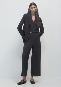 Completo tailoring nero con maniche lunghe, un gilet corto e pantaloni a gamba larga. Tessuto liscio, design minimalista e dettagli dei bottoni discreti.