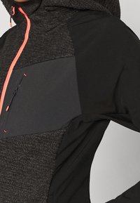 Schwarze Mehrtextur-Jacke mit figurbetontem Design, ausgestattet mit einer Reißverschlusstasche, einem leuchtend orangefarbenen Reißverschluss und einer Kapuze. Der Stoff kombiniert glatte und gemusterte Texturen.