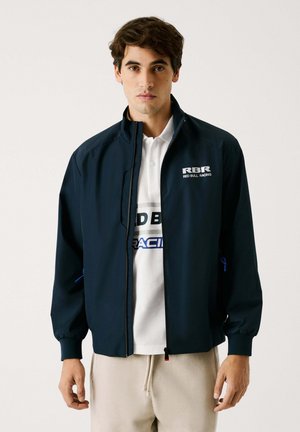 Navy windbreaker jacket with a front zipper, elastic cuffs, and Red Bull Racing logo. Worn over a white shirt with blue accents.