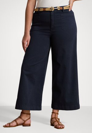 Pantalons larges couleur marine en coton, avec une texture lisse, des poches arrière et une ceinture tressée beige, associés à des sandales brunes à lanières.