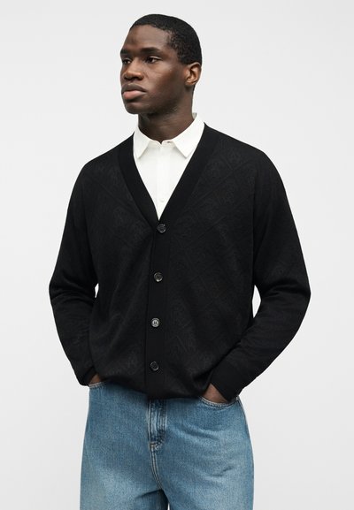 Άνδρας με μαύρη κουμπωτή ζακέτα με σχέδια πάνω σε λευκό πουκάμισο με γιακά, με τα χέρια στις τσέπες ανοιχτόχρωμων τζιν, κοιτάζει ελαφρώς προς τα αριστερά.