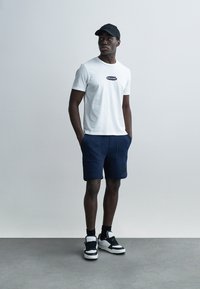 Λευκό T-shirt με μαύρο λογότυπο, σκούρο μπλε σόρτς και μαύρο καπέλο. Ολοκληρωμένο με μαύρα και λευκά αθλητικά παπούτσια. Μινιμαλιστικός σχεδιασμός, casual εμφάνιση.