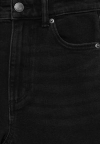 Gros plan sur un jean en denim noir montrant la poche avant, la passants de ceinture, un bouton en métal, et des rivets avec des détails de couture visibles.