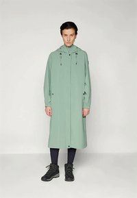 Άτομο που στέκεται φορώντας ένα μακρύ, ανοιχτό πράσινο πανωφόρι με κουκούλα, πάνω από μαύρες κολάν και μαύρα παπούτσια πεζοπορίας, σε λευκό φόντο.