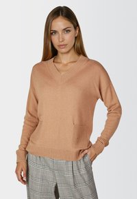 V-neck, light brown sweater made of soft material with ribbed cuffs, two front pockets, and a relaxed fit. Paired with patterned trousers.