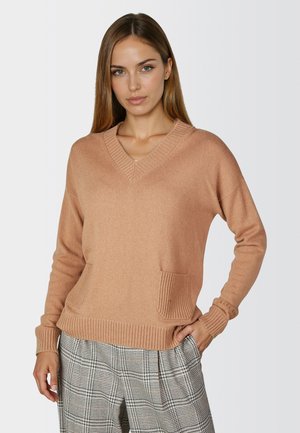 V-neck, light brown sweater made of soft material with ribbed cuffs, two front pockets, and a relaxed fit. Paired with patterned trousers.