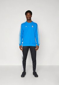 Blå tröja med vita ränder och logga, kombinerad med svarta byxor med vita randdetaljer och svarta sportskor.