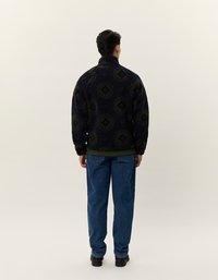 Marinefarget fleecepullover med grønne geometriske mønstre, høy krage og elastisk kant. Kombinert med blå jeans og svarte sko.