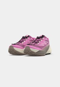 Chaussures de trail running roses avec une tige en mesh, des lacets noirs et une semelle externe texturée marron. Elles présentent un intersemelle blanche rembourrée pour un meilleur soutien.