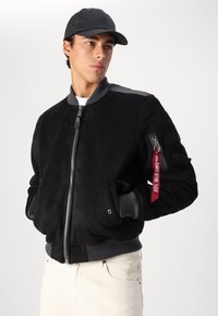 Giacca bomber nera in suede, con polsini e orlo a coste, chiusura frontale con zip, tasche laterali e tasca con zip sulla manica con etichetta rossa.