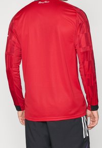 Maglietta sportiva rossa a maniche lunghe con un design testurizzato e dettagli neri sui polsini e ai lati. Tessuto liscio con una forma aderente.