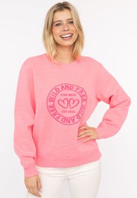 Rosa Sweatshirt mit Rundhalsausschnitt, langen Ärmeln und gerippten Bündchen. Enthält ein kreisförmiges Grafikdesign mit dem Text "STAY WILD" und Herzen in der Mitte.