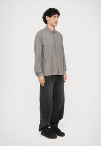 Camicia button-up grigia con motivo a righe verticali; abbinata a pantaloni in denim neri larghi con dettagli di cucitura laterale e sneakers nere.