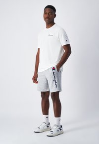 Weißes T-Shirt mit kurzen Ärmeln und kleinem Logo, kombiniert mit grauen Sweatshorts mit einem dunkelblauen Logo-Print. Trägt dazu weiße Sneaker und Socken.