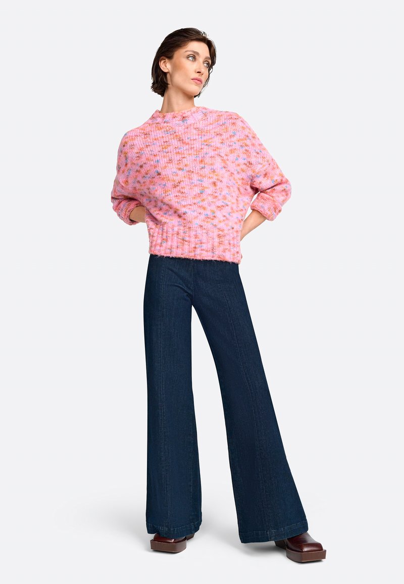 Rosa gestrickter Pullover mit hohem Kragen und strukturiertem Stoff, kombiniert mit dunkelblauen Schlagjeans und braunen Plateauschuhen.