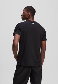 Zwarte katoenen T-shirt met korte mouwen, met een ronde hals en een klein logo op de achterkant. Draagt met zwarte broek.