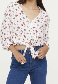 Blusa blanca de botones con un patrón floral rojo y azul, atada por delante, con escote en V y mangas sueltas, combinada con jeans azules.