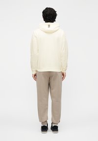 Jasnokremowa bluza z kapturem ze ściągaczem i paskiem z tyłu, zestawiona z luźnymi beżowymi spodniami dresowymi i czarnymi butami sportowymi.