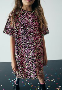 Svart kortärmad klänning med spridda rosa och gröna blommönster. Modellens protesben är synligt, med skinande silverdetalj. Konfetti i bakgrunden.