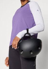 Lila texturerad långärmad topp med vita ärmar och en svart hjälm med ventilationshål. Hjälmen har ett rem för säker fastsättning.