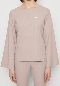 Dlouhý rukáv, žebrovaný top ve světle růžové barvě, s kulatým výstřihem a malým bílým logem Nike na hrudi. Boční rozparky na lemu.