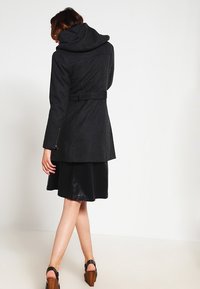 Abrigo negro de lana con capucha, cintura ceñida con cinturón y mangas largas. Presenta bolsillos laterales con cremallera y un corte entallado, combinado con una falda negra.