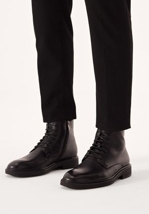 Botas negras de cuero hasta el tobillo con cordones y cremallera lateral, con punta redonda y suela plana, combinadas con pantalones negros de corte ajustado.