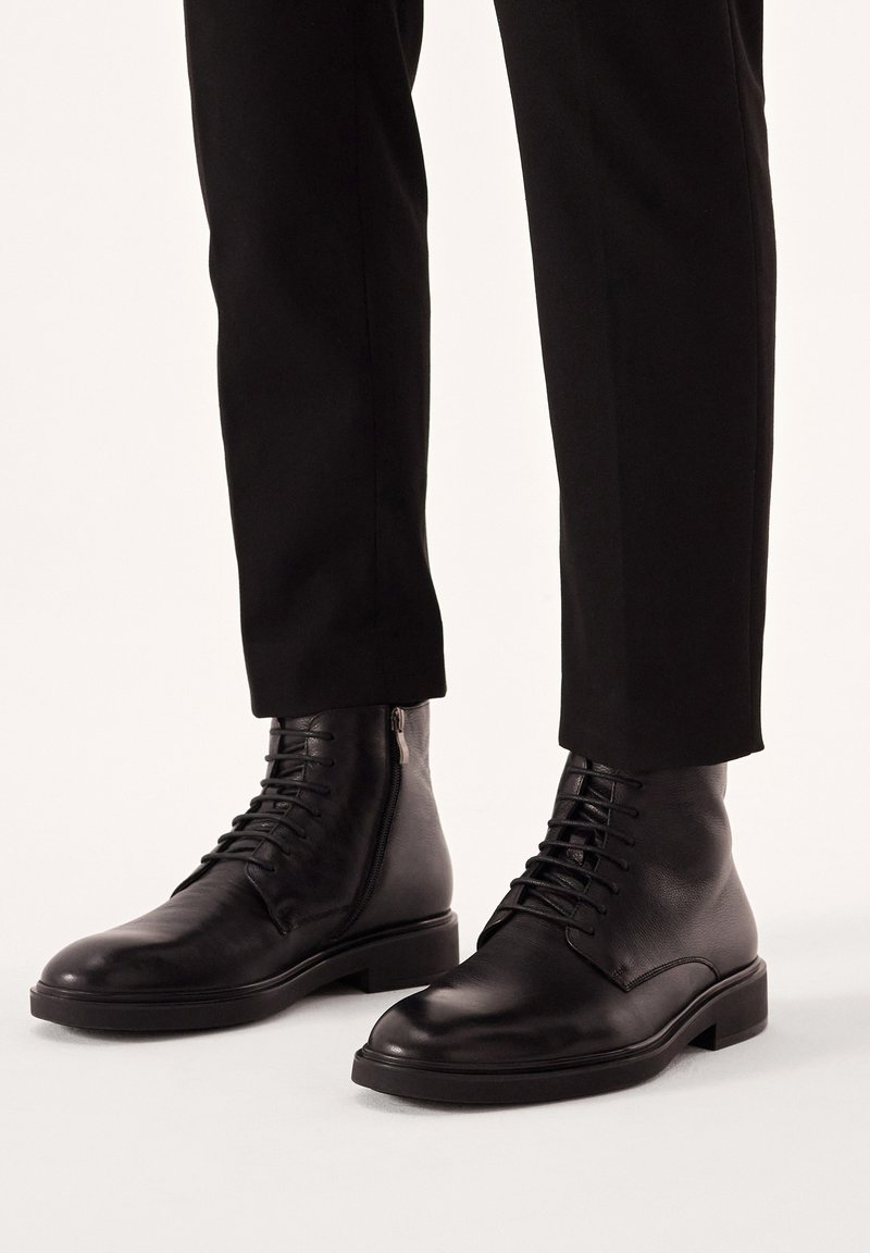 Botas negras de cuero hasta el tobillo con cordones y cremallera lateral, con punta redonda y suela plana, combinadas con pantalones negros de corte ajustado.