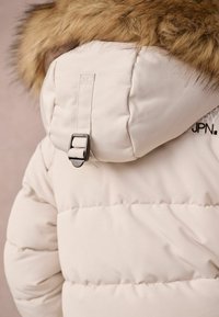 Puffermantel in hellbeige mit abnehmbarem Fellkapuze. Verfügt über eine Metallspange und ein gesticktes Logodetail auf der rechten Seite.